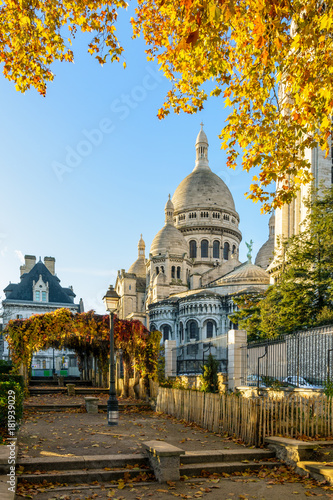 Rear view of the Basilica of the Sacred Heart of Paris in autumn at sunrise seen from a public park with orange leaves in the foreground against blue sky.