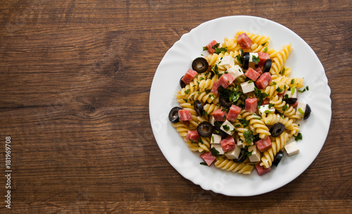 pasta fusilli salad with salami, cheese and olive in white plate on a wooden background. with copy space. top view