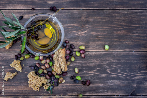 Bowl with extra virgin olive oil, olives, a fresh branch of olive tree and cretan rusk dakos close up on wooden table, Crete, Greece.