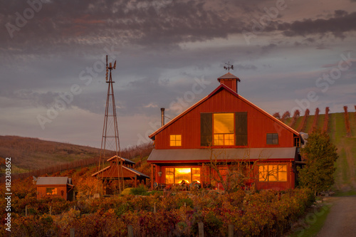 A red house with a windmill at sunset is set in a vineyard with autumn colors. The sunset is reflected in the windows of the house. A blue sky with clouds are in the background.