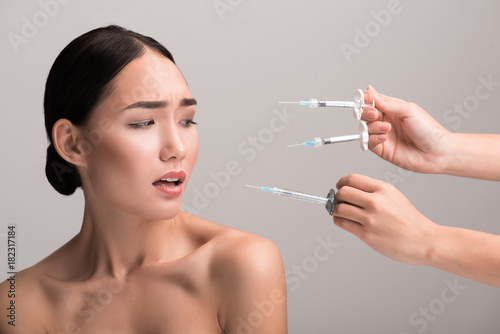 Gorgeous young naked asian woman is scaring of botox injections. She is looking with fear at syringes in hands of doctors near her face. Isolated background. Anti-aging skincare concept