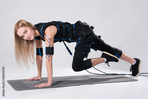 Beautiful blonde in an electric muscular suit for stimulation makes an exercise on the rug