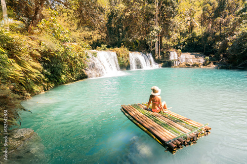 Woman in bikini and hat sitting on bamboo raft and enjoying view on waterfall.