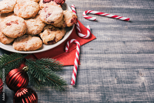 Traditional British dessert, scones with strawberries. Sweet coconut cookies with raspberry jam and yogurt on wooden table. Candy cane, fir-tree branches, baubles, cup of milk