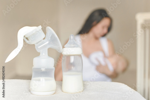 Manual breast pump and bottle with breast milk on the background of mother holding in her hands and breastfeeding baby. Maternity and baby care.