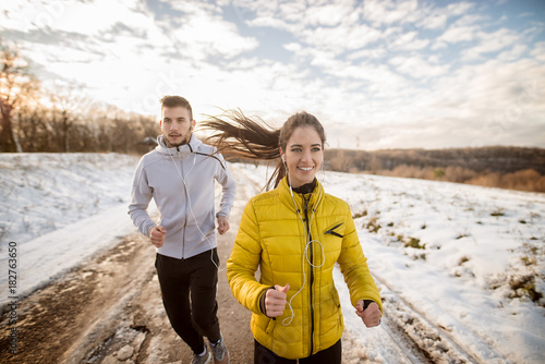 Charming young active runner girl jogging with her personal fit trainer on a snowy road in nature.