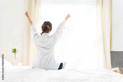 Back view of woman stretching in morning after waking up on bed near window. Holiday and Relax concept. Lazy day and Working day concept. Office woman and worker in daily life theme
