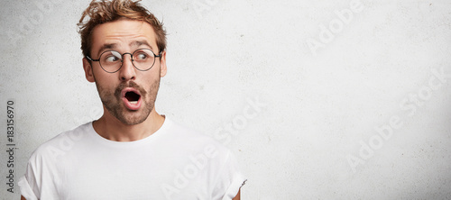 Horizontal isolated shot of surprised male college teacher, wears spectacles, wonders that he should work on weekends, doesn`t expect to hear about expel of clever student, poses against concrete wall