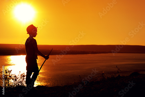 Silhouette of little boy in hat with stick looking far into the sea in warm sunset evening
