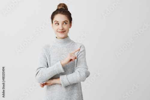 Pretty brunette woman posing on camera with tricky look and gesturing to side. Female designer presenting lovely product with index finger. Gesture concept