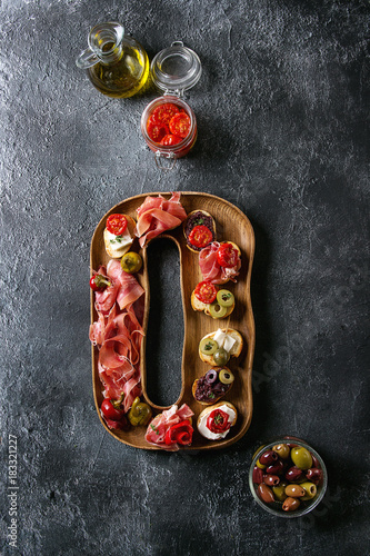 Tapas or bruschetta variety. Crusty bread with ham prosciutto, sun dried tomatoes, olive oil, olives, pepper on decorative wooden plate over dark texture background. Top view with space
