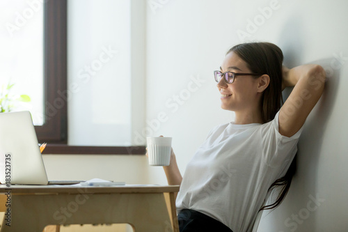 Satisfied young businesswoman holding hand behind head and drinking coffee. Beautiful female manager having break, resting and smiling