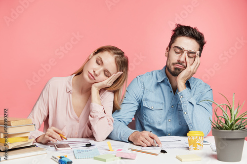 Portrait tired crew of office workers sit at table, fall asleep after working long hours on preparing startup project, feel tiredness, isolated over pink background. People and overworking concept