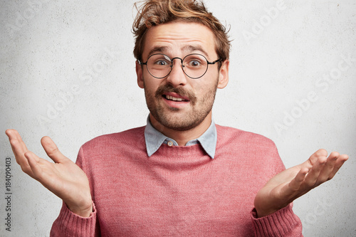 Clueless doubtful male wonk wears round spectacles, shrugs shoulders in bewilderment as doesn`t know answer on question, hesitates, isolated over white concrete background. Body language concept