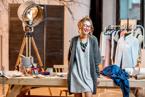 Portrait of a young female fashion designer standing at the office with different tailoring tools and clothes