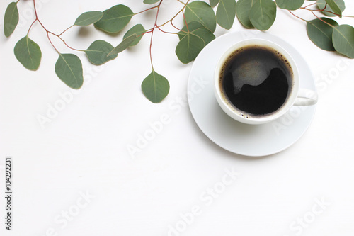 Floral composition made of green eucalyptus leaves and branches on white wooden background with cup of coffee. Feminine office desk, styled stock image, flat lay, top view with empty space.