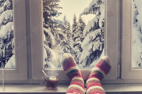 feet in wool knitted socks and a cup of steaming tea on the windowsill overlooking the winter snow forest through the window