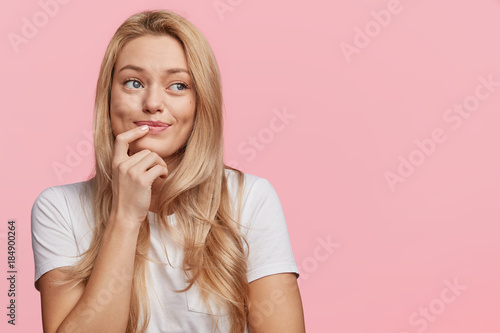 Pretty lovely blonde female with pure healthy skin dressed casually looks with dreamy expression aside, remembers pleasant moments, isolated over pink background, copy space for your advertisment