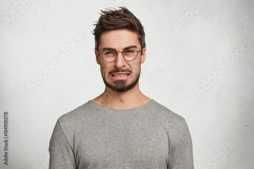 Portrait of handsome bearded male with disgusting expression sees sour lemon in front, frowns in displeasure, isolated over white concrete background. Male expresses aversion towards something