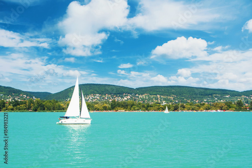 Panoramic view of lake river blue transparent water, a white sport modern luxury yacht sail boat floating and a green shore with forest, hills, villages and beach. Holiday by the water and sail race.