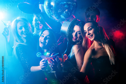 Dance party with group people dancing. Women and men have fun in night club. Happy girl on foreground and disco ball on background. Illumination from rays, special effects. Opening of new strip club.