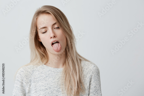 Negative human reaction, feelings and attitude. Portrait of bored annoyed blonde girl in casual wear grimacing, sticking out her tongue, blinking, feeling nauseous because of bad smell or stink