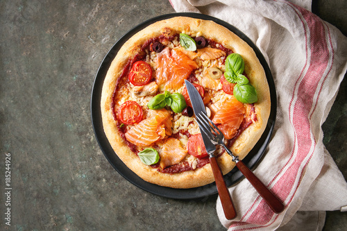 Traditional pizza with smoked salmon, cheese, tomatoes and basil served on black plate with kitchen towel over old dark metal background. Top view with space. Rustic style