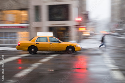 Panning motion image of a New York City yellow taxi cab in the snow as it passes through an intersection and past a pedestrian