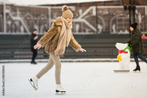 Full length portrait of young female with blonde hair in fur coat, beige hat, scarf and trousers skating on ice rink, outdoors at winter /Weekends activities outdoor in cold weather/