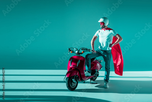 man in protective helmet, superhero mask and cape standing at red scooter