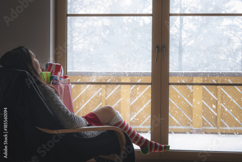 Woman in armchair in front of a snowy window - Young brunette woman wearing a cozy sweater and long red and white socks, sitting on an armchair in front of big glass door looking at the snowfall.