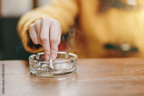 Focus on caucasian young woman hand putting out cigarette on glass ashtray on wooden table, cigarette butt, smoking is dying. Quit smoking. Health concept. Close up photo.