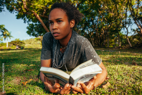 Beautiful young woman with afro hairstyle and book in her hands lying on the grass in the park on sunny summer day