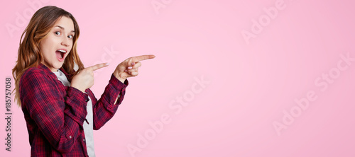 Excited beautiful woman stands sideways, wears chemise, points with fore fingers aside at blank copy space for your advertisment or promotional text. Joyful female model poses in pink studio.
