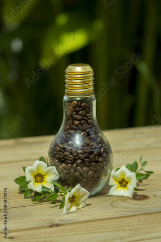 a pile of brown coffee beans with a small jar of glass bulb shape with more coffee beans on wooden table with available light at the outdoor