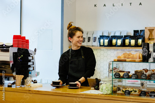 Friendly hospitable woman barista holding a two coffee cups with smiling face at cafe counter background, food and drink industry concept. Modern coffee shop interior.