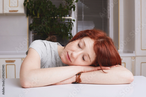 Tired ginger housewife takes nap, leans head on hands, being fatigue after cleaning kitchen, closes eyes, sits against kitchen interior. Overworked female student falls asleep. People, work, tiredness