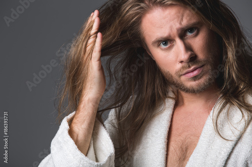 Portrait of displeased bearded guy holding his long locks among fingers and looking at the camera. Isolated on grey background