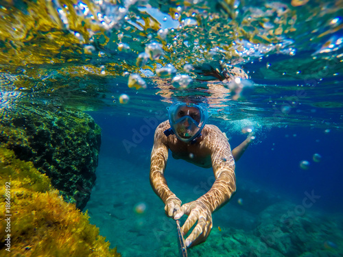 Underwater view of a young diver man swimming in the turquoise sea under the surface with snorkelling mask for summer vacation while taking a selfie with a stick.