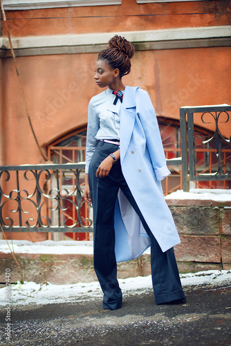 African girl with bright makeup and a blue coat stands and looks to the side on the snowy streets in style fashion