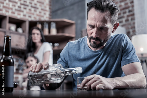 Drinking father. Serious unhappy sad man sitting at the table and pouring vodka into his glass while having alcohol addiction