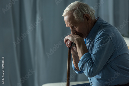 Senior sad man leans on a cane while sitting on sofa