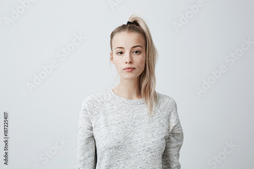 Attractive beautiful female with blonde pony tail, feeling self-assuarance while posing against blank studio wall. Confident woman with dark eyes dressed casually isolated against gray background