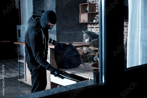 Professional burglar. Violent professional masked burglar wearing a black uniform and gloves and stealing a laptop from the table and holding a bag