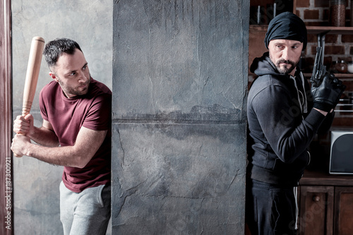 Fighting. Determined dark-haired beaded man holding a stick and hiding from the killer standing behind the wall and holding a gun