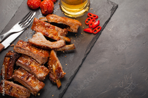 Pork ribs in barbecue sauce and honey baked tomatoes and a glass of beer on a black slate dish. A great snack to beer on a dark stone background. Top view with copy space