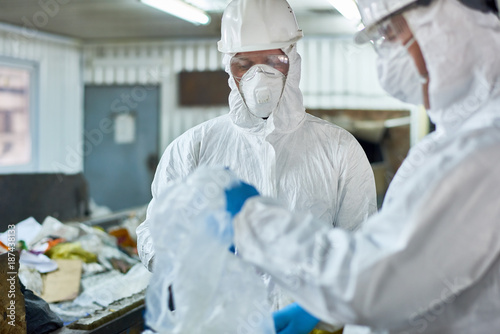 Portrait of two workers wearing biohazard suits sorting recyclable plastic and cardboard at waste processing plant