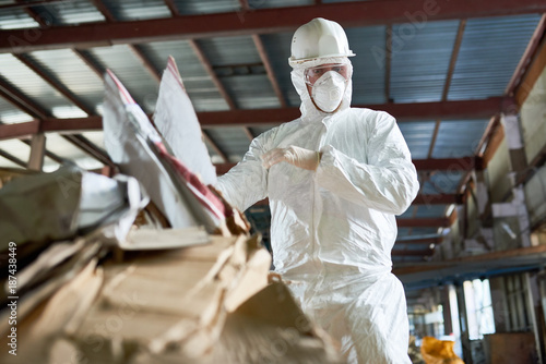 Low angle portrait of factory worker wearing biohazard suit sorting reusable cardboard on waste processing plant, copy space