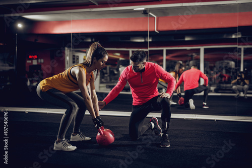 Young healthy focused sporty flexible shape girl with a ponytail doing squat exercises with the kettlebell while handsome helpful personal trainer crouching next to her.