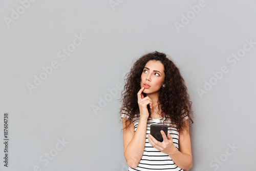 Picture of nice woman touching her lips and looking aside while thinking about something important or trying to remember over grey background